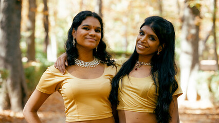 Two young indian women posing for a portrait, wearing traditional jewelry and gold tops, embracing...