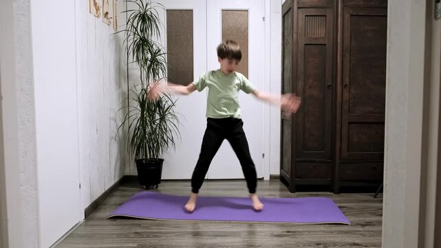 Caucasian boy doing an exercise Jumping Jacks on yoga mat at home. Children workout. Morning warmup. Child strengthening the muscles of legs, endurance and balance. Strong sporty body. HQ 4k footage.