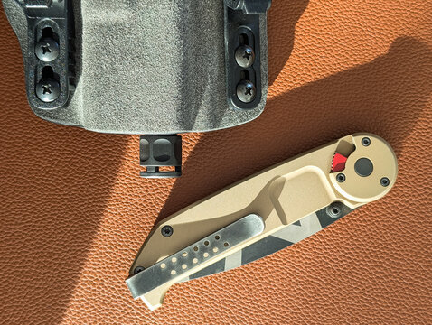 Close up of tactical folding knife and handgun with compensator in holster on brown leather surface, everyday carry EDC gear