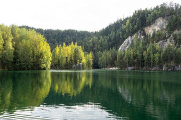 Fototapeta premium Calm forest lake reflecting dense trees and rocky slopes in green water. Gentle ripples spread across the surface creating a peaceful natural landscape scene.