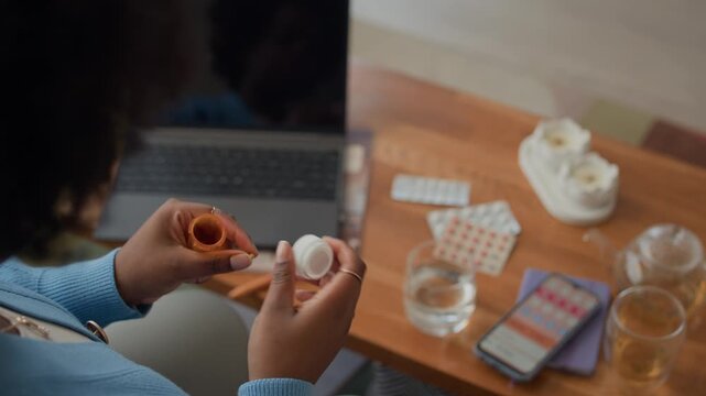 Over the shoulder view of adult Black woman taking contraceptive pills and checking ovulation calendar in app on smartphone at home