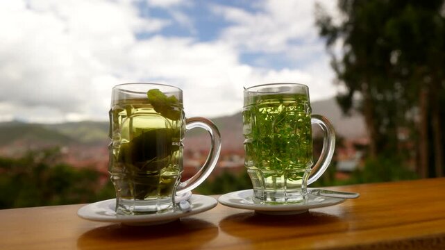 Glass mugs of mate coca and muna tea drinks on table with Cusco city and Andes in background, hand grabs single drink away