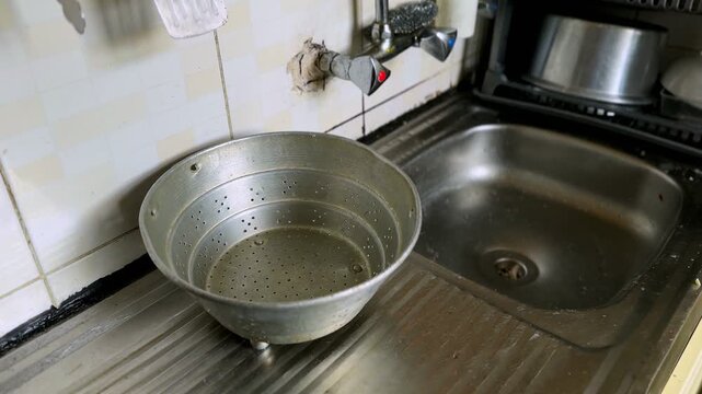 Old perforated metal colander placed on a stainless steel drainboard next to a kitchen sink with wall mounted faucet and stained tiled walls in natural indoor lighting.