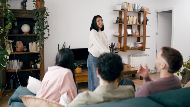 Four young adult friends playing an acting game in a cozy living room. A woman is gesturing while her companions laugh and guess from the sofa