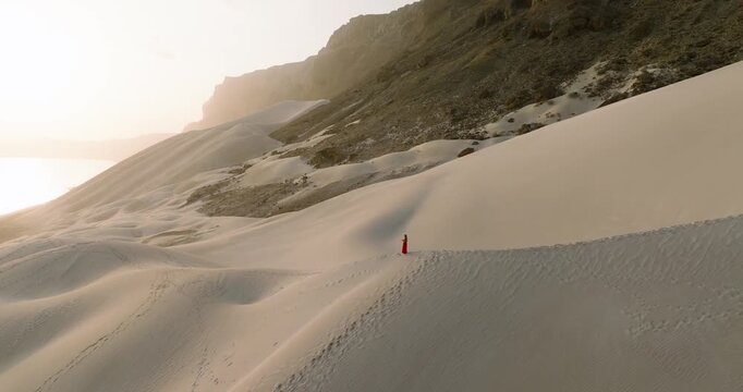 Aerial view of a person in red standing on the crest of a vast, undulating sand dune, contrasting with the rugged mountain backdrop, Arher, Socotra.