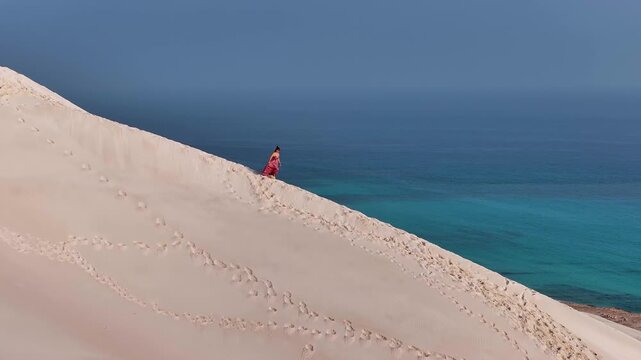 Aerial view of a woman in a pink dress walking on the sand dune near the turquoise sea, Socotra, Socotra.