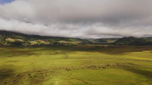 Aerial view of Phobjikha Valley, a vast green expanse cut by a winding stream beneath a sky heavy with clouds, creating a tranquil, Bhutan.