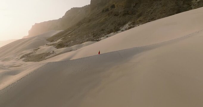 Aerial view of a woman in a red dress traversing the vast, undulating sand dunes against the backdrop of the rugged cliffs, Arher, Socotra.