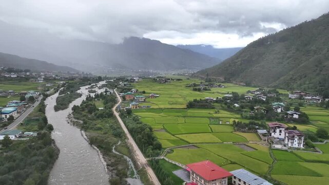 Aerial view of a river snaking through a valley with bright green fields contrasting against the cloudy sky and distant mountains, Paro, Bhutan.