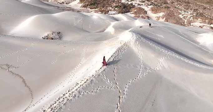 Aerial view of the pristine white sand dunes meeting the rugged mountain terrain, where a woman in pink walks amidst countless footprints, Socotra Island, Socotra.
