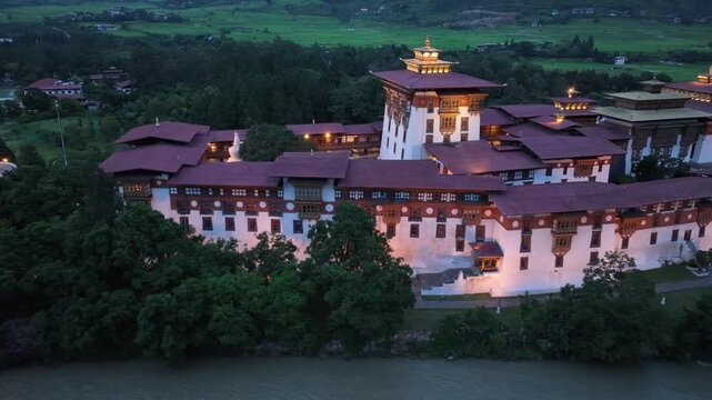 Aerial view of majestic Punakha Dzong, its white walls and red roofs contrasting with the dark green trees and the river flowing by, Punakha, Punakha, Bhutan.