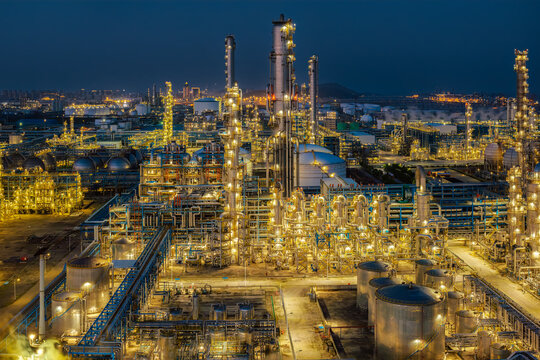 Massive industrial oil refinery and chemical processing plant illuminated at night with complex piping, storage tanks, and tall distillation towers.