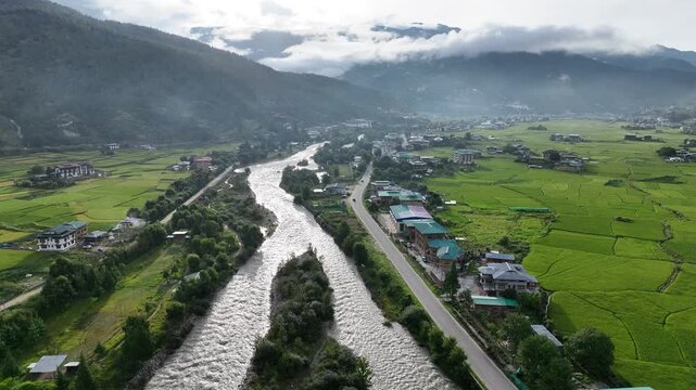 Aerial view of a river flowing through vibrant green rice fields, a road running parallel, with mountains in the background, Paro, Bhutan.