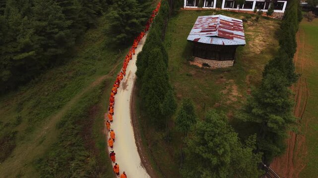 Nubding, Bhutan - 21 September 2025: Aerial view of monks in orange robes walking along a path near buildings and green trees under a cloudy sky.