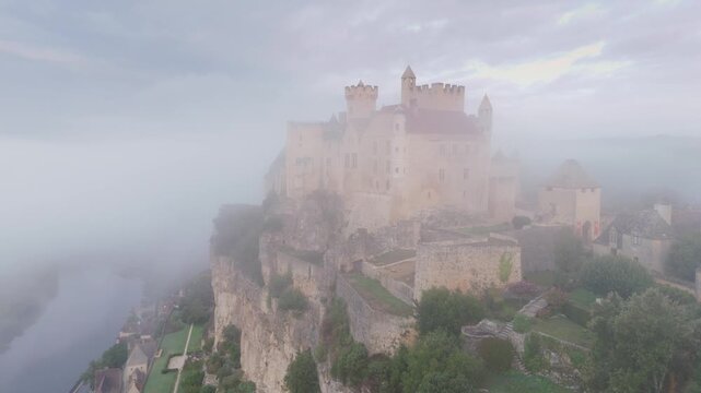 Beynac-et-Cazenac, France - 12 September 2025: Aerial view of Chateau de Beynac perched atop a cliff, shrouded in mist, overlooking a tranquil river below.