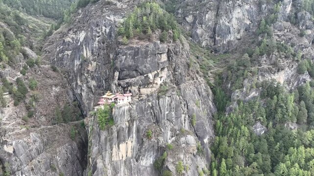 Aerial view of Tiger's Nest Monastery dramatically clinging to a cliff, surrounded by lush greenery and rugged terrain, Taktsang, Paro, Bhutan.