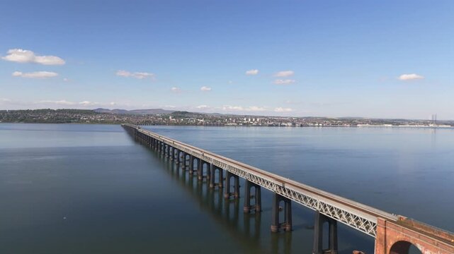 Aerial view of the Tay Rail Bridge spans the tranquil water, connecting the land under a blue sky with scattered clouds, Dundee, United Kingdom.