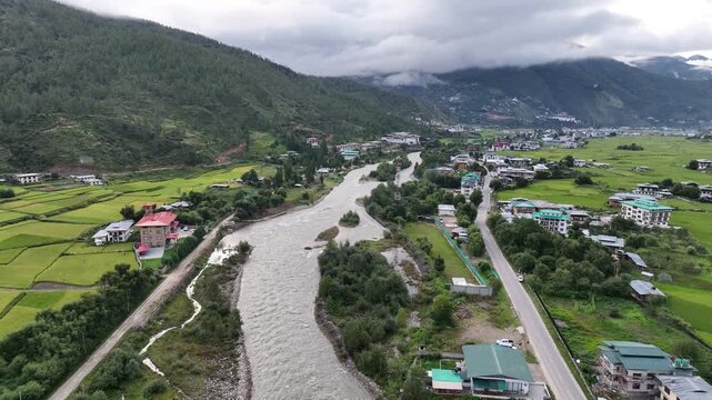 Aerial view of a river meandering through the lush green valley, flanked by rice paddies and buildings, under a cloudy sky, Paro, Bhutan.