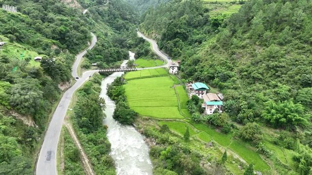 Aerial view of bright green rice fields and traditional houses nestled between hills, bisected by a river and roads, Whachey Lhakhang valley, Bhutan.