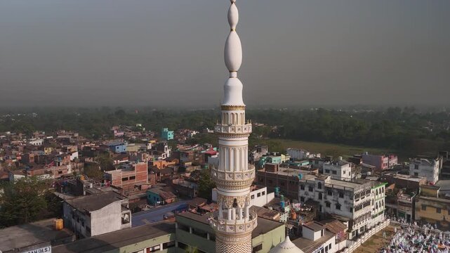Aerial view of a tall white minaret rising above the cityscape, contrasting with the dense buildings and green spaces, Malda, West Bengal, India.