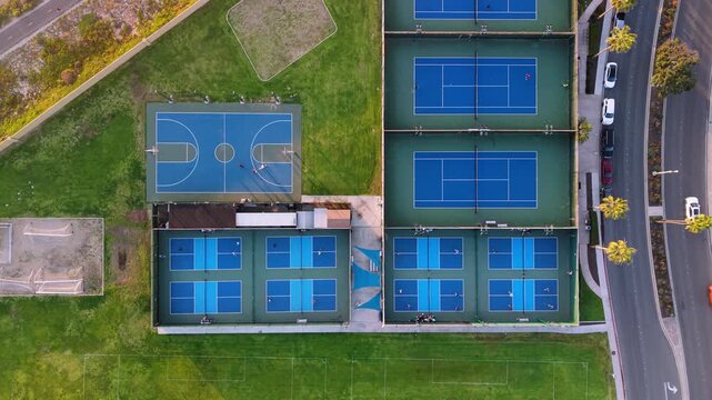 Aerial view of blue tennis courts, a basketball court, and a road with parked cars surrounded by lush green grass, Coronado, California, United States.