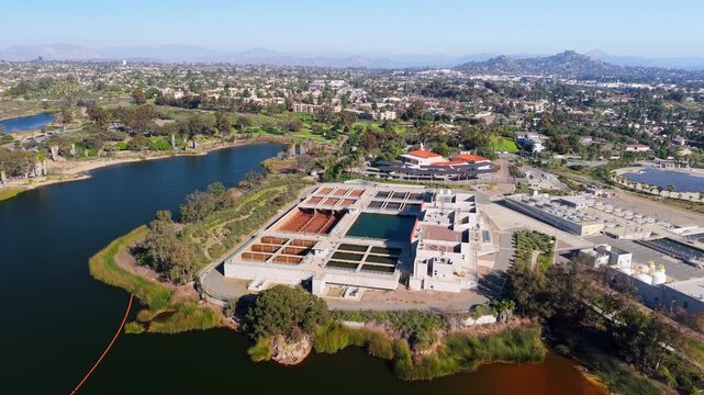 Aerial view of a water treatment plant along a dark lake, reflecting the sky, contrasted by lush green trees, La Mesa, California, United States.