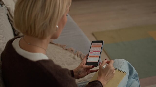 Over the shoulder view of Caucasian middle-aged blonde woman counting cycle days while using application on smartphone and taking notes at home