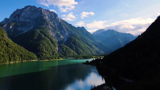 Aerial view of the lake with mountains surrounded by green trees, creating a scenic landscape with contrasting light and shadows, Friuli-Venezia Giulia, Italy.