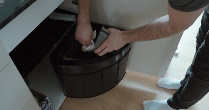 Person emptying leftover food from a container into a kitchen waste bin before placing dishes into the dishwasher, emphasizing hygiene, proper cleaning routine, and household efficiency.