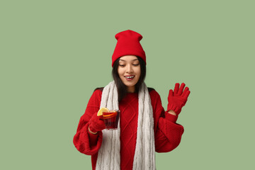 Beautiful young happy Asian woman in winter clothes with cup of hot mulled wine waving hand on...