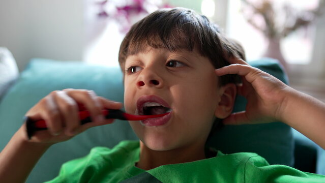 Young boy brushing teeth with red toothbrush, mouth open, toothpaste foam visible, sitting on couch at home, relaxed posture, hand on head, natural window light