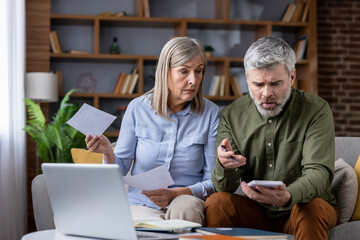 Senior couple sitting on a couch, focused on paperwork and a calculator, discussing bills and tax...