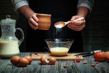 A person pours sugar from a container into a bowl with milk and eggs on a wooden surface. The setting is a kitchen with dim lighting and various kitchen items nearby © Віталій Б.