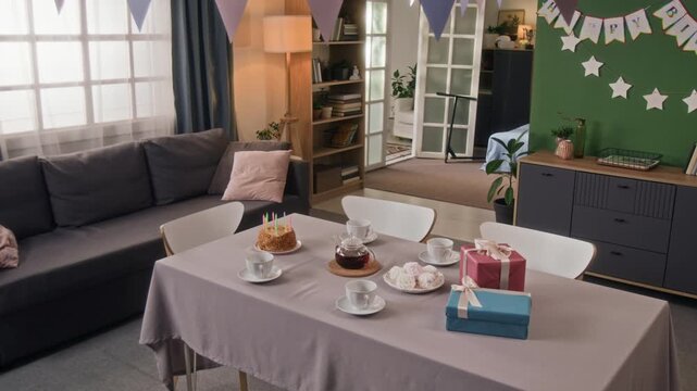 No people zoom in view of interior of living room decorated with garlands featuring dining table set for tea party with birthday cake, four cups, two gift boxes, teapot and marshmallows on plate