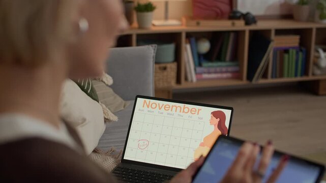 Zoom in shot of middle-aged Caucasian woman using application for meditation on digital tablet while sitting on sofa with pregnancy calendar on laptop screen in living room