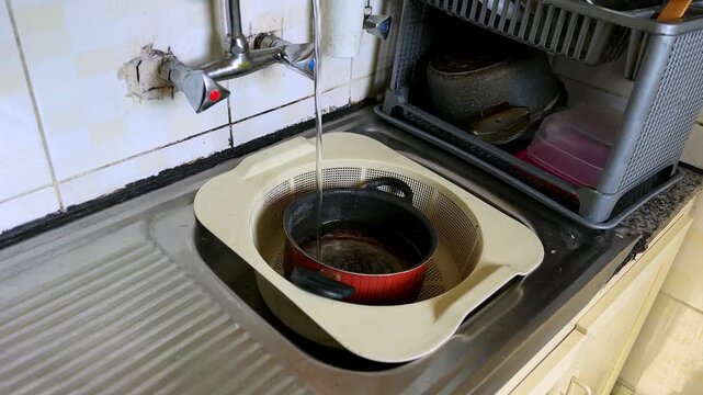 Water flowing from a wall mounted faucet into a red cooking pot placed inside a plastic colander in a worn kitchen sink with tiled walls and dish rack in natural indoor light.