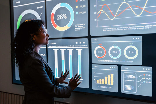 Professional analyzing an interactive data dashboard with charts and graphs on a large screen, reviewing metrics and trends in a modern office setting.