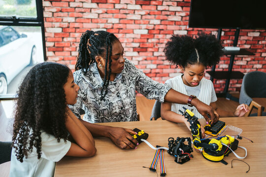Teacher guides two girls building a robotic arm kit in a classroom makerspace, using electronics and tools for hands-on STEM learning