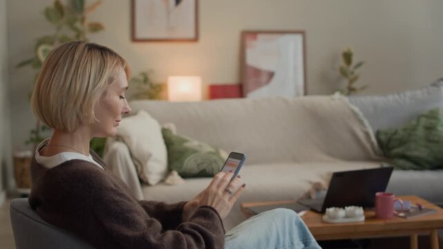 Side view of adult Caucasian woman tracking cycle in app on smartphone while relaxing in chair in cozy living room