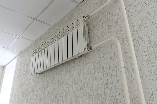 A white bimetallic radiator is mounted on a textured light grey wall, connected to white plastic pipes running vertically and horizontally; set against a tiled ceiling