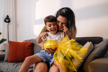 Brazilian mother surprising young son with handmade Easter egg gift on living room sofa,...