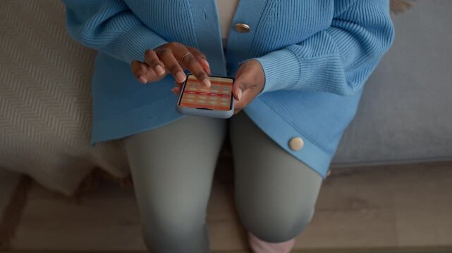 Top down shot of unrecognizable woman checking smartphone with menstrual calendar while sitting by table with pills and tea at home