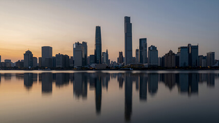 Obraz premium Serene City Skyline at Dusk with Modern Skyscrapers Mirrored in Calm Water Reflecting Warm Sunset Glow over Urban Landscape