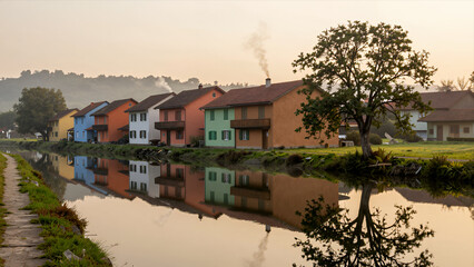 Obraz premium Serene Row of Colorful Houses Reflected in Calm Canal Water during Golden Hour Sunrise or Sunset with Misty Atmosphere and Tree Silhouette