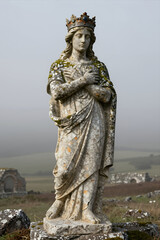 Fototapeta premium Weathered Stone Statue of a Crowned Woman with Draped Robe Standing in Misty Rural Landscape, Ancient Monument, Historical Sculpture Outdoors