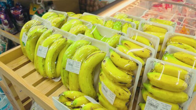 Bananas individually packaged in plastic cling film and white foam trays, arranged in rows on a wooden display stand inside a supermarket, representing consumer convenience and food merchandising