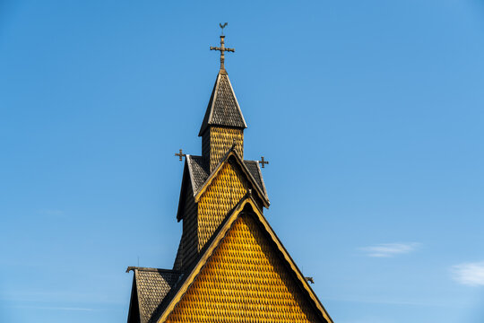 View of a wooden Heddal Stave church with a cross on top stands silhouetted against a clear blue sky, its intricate details highlighted by the sun, Heddal, Telemark, Norway.