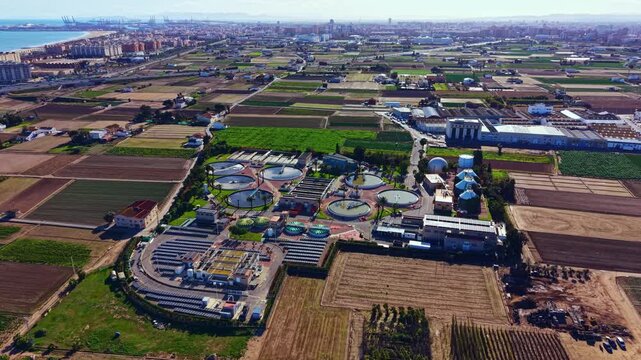 A waste treatment facility is situated in a rural area surrounded by agricultural fields. The facility has circular tanks and green spaces. A city and ocean are visible in the distance.