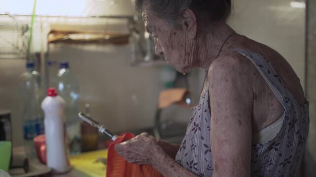 Senior elderly woman washing dishes in kitchen. Sad mature housewife cleans up the old dishes in the kitchen. Upset woman with gray hair 90 years old washing kitchen utensils in a hand wash at home.