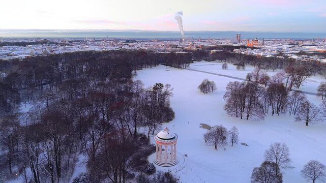 Winter sunrise over Munich old town skyline. Aerial drone view from the English Garden showing the snow-covered Monopteros and historic city center landmarks like Frauenkirche and Town Hall